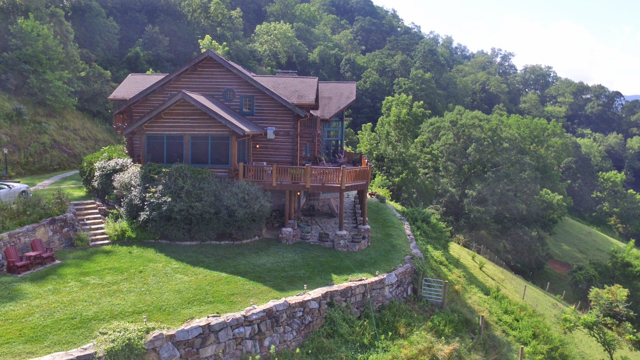 West side view of log lodge with rockers in lawn and side view of main deck and rock patio. The entrance walking path is to the left and the boulder retaining wall is on the right of the log lodge.