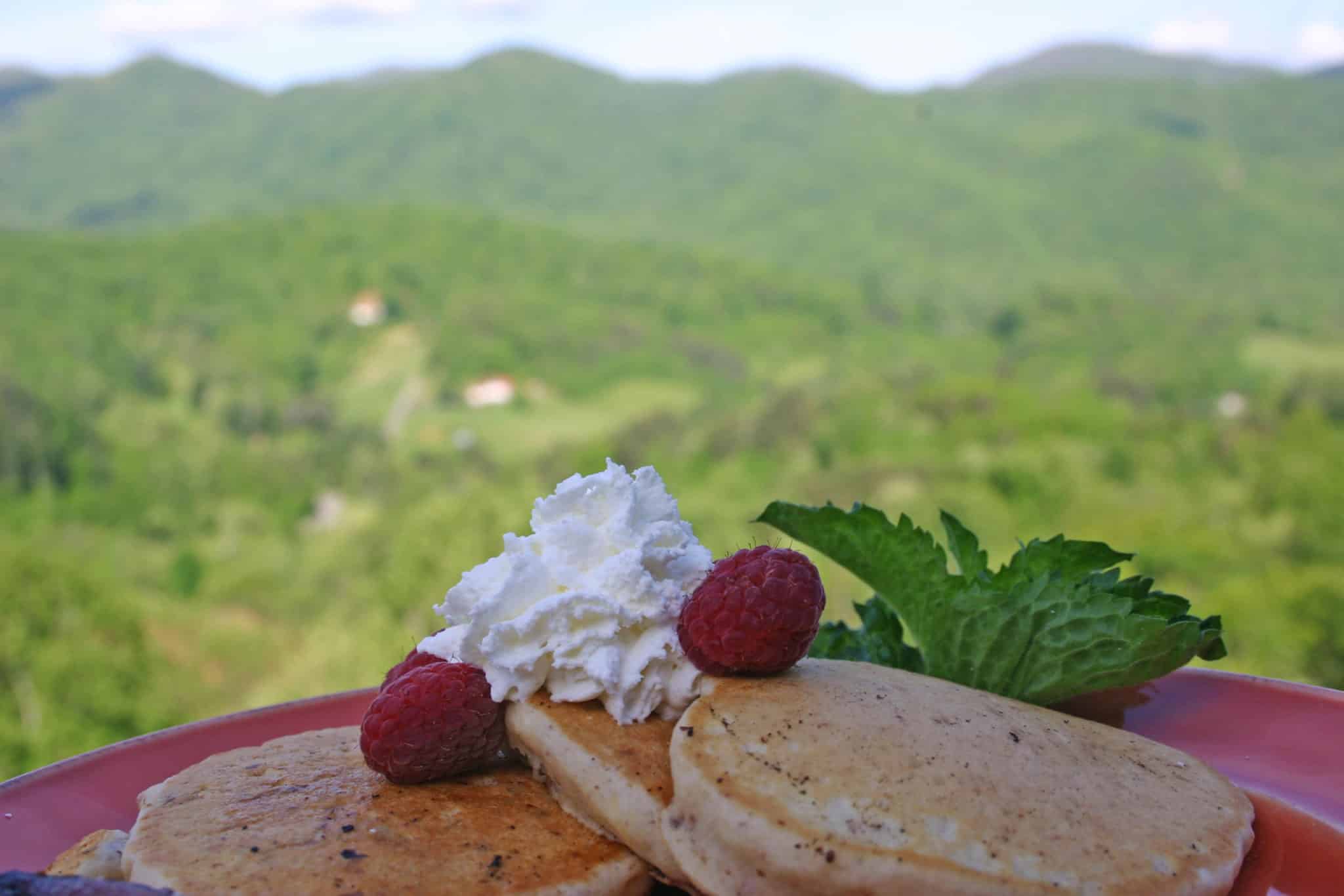 Three pancakes overlapping on red plate. Topped with two raspberries and whipped cream. Green hills in background.