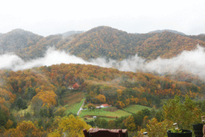 Green grass in valley around a red roofed home. Mountains in distance with orange and green and clouds hanging over the first ridge.
