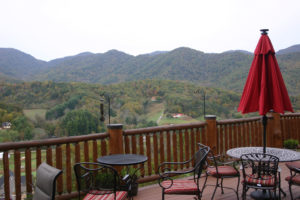 Mountains in distance that are still mostly green spotted with yellow and red fall foliage. foreground is deck with red umbrella and black iron table.