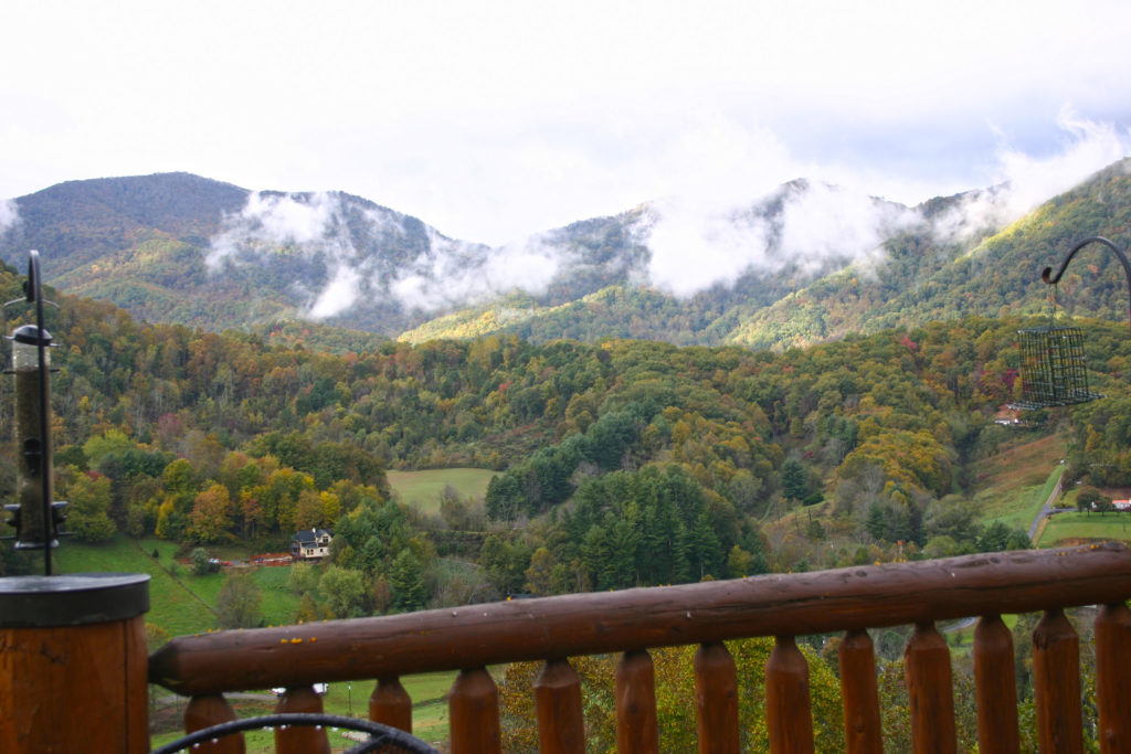 Puffy clouds in front of mountains with fall foliage colors of red, orange and yellow. Deck railing in foreground