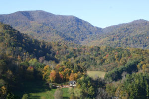 house across valley with red and orange leaved trees in side yard. behind the house are mountains with similiar fall colors mixed with leaves that arte still green.