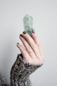  Woman holding green quartz pointed up with her fingertips. Brown and white sweater covers hand. Short nails painted dark. Wearing thin gold ring.