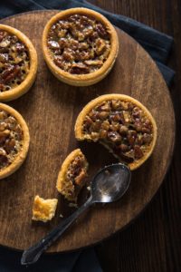 Wooden round tray holding 4 pecan tarts, 2 half visible, 1 cut into with black spoon.