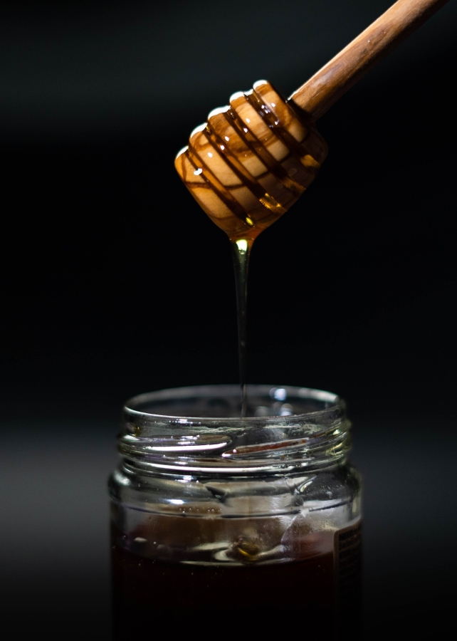 Honey dipper several inches above shallow jar of honey against black background