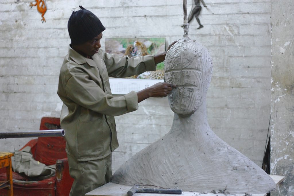 Man in skull cap and beige clothes sculpting face of larger-than-life bust in white-bricked studio.