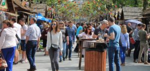 Street fair with people walking, eating at tables, stopped at booths. Colorful decorations strung across street 