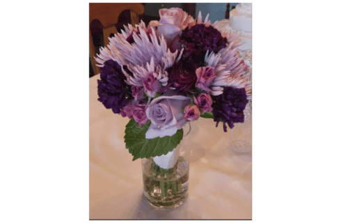 A bouquet of dark purple and lavendar daisies and other flowers with green leaves in a glass vase as a bride's bouquet