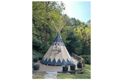 A Native American TiPi sits in front of a blue sky and green trees