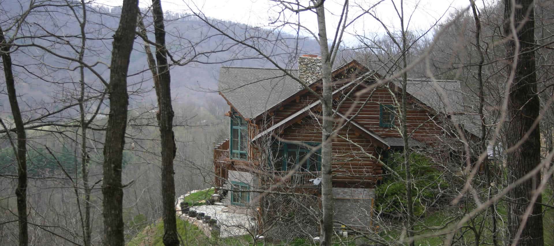Side view through trees of large log-cabin home with a stone patio and stone lower level.