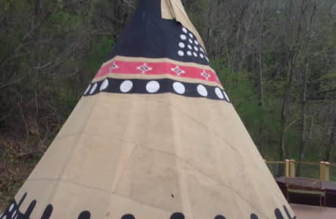 Beige and black with red tipi on a wooden deck next to woods.