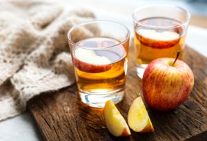 2 glass, short tumblers (old-fashioneds) with cider with apple slice in glass. Plus apple and apple slices next to glass on wooden tabletop.