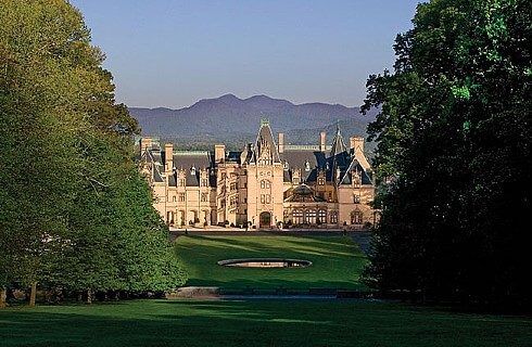 Large light stone colored estate with large green front lawn and tall trees with mountains in the distance
