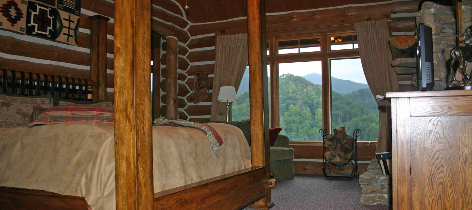 Large wooden poster bed in cozy bedroom with large windows overlooking tree-covered hills.