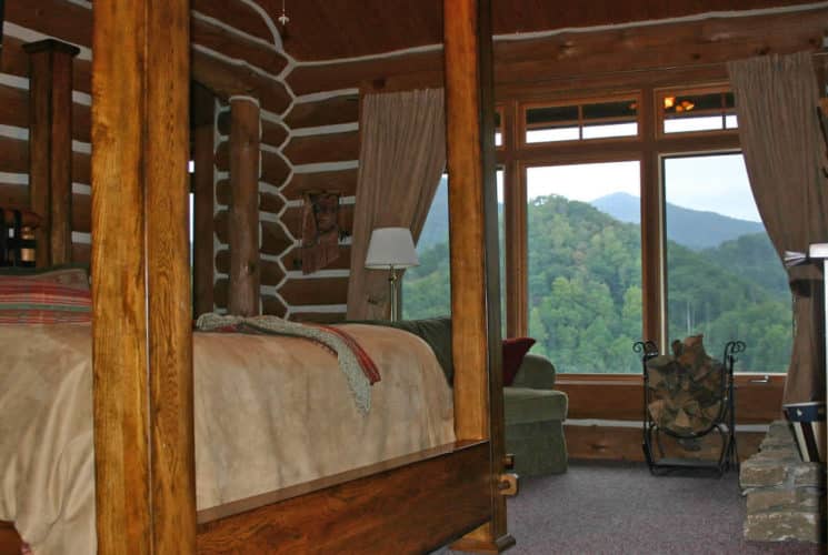 Large wooden poster bed in cozy log-cabin bedroom with large windows overlooking tree-covered hills.