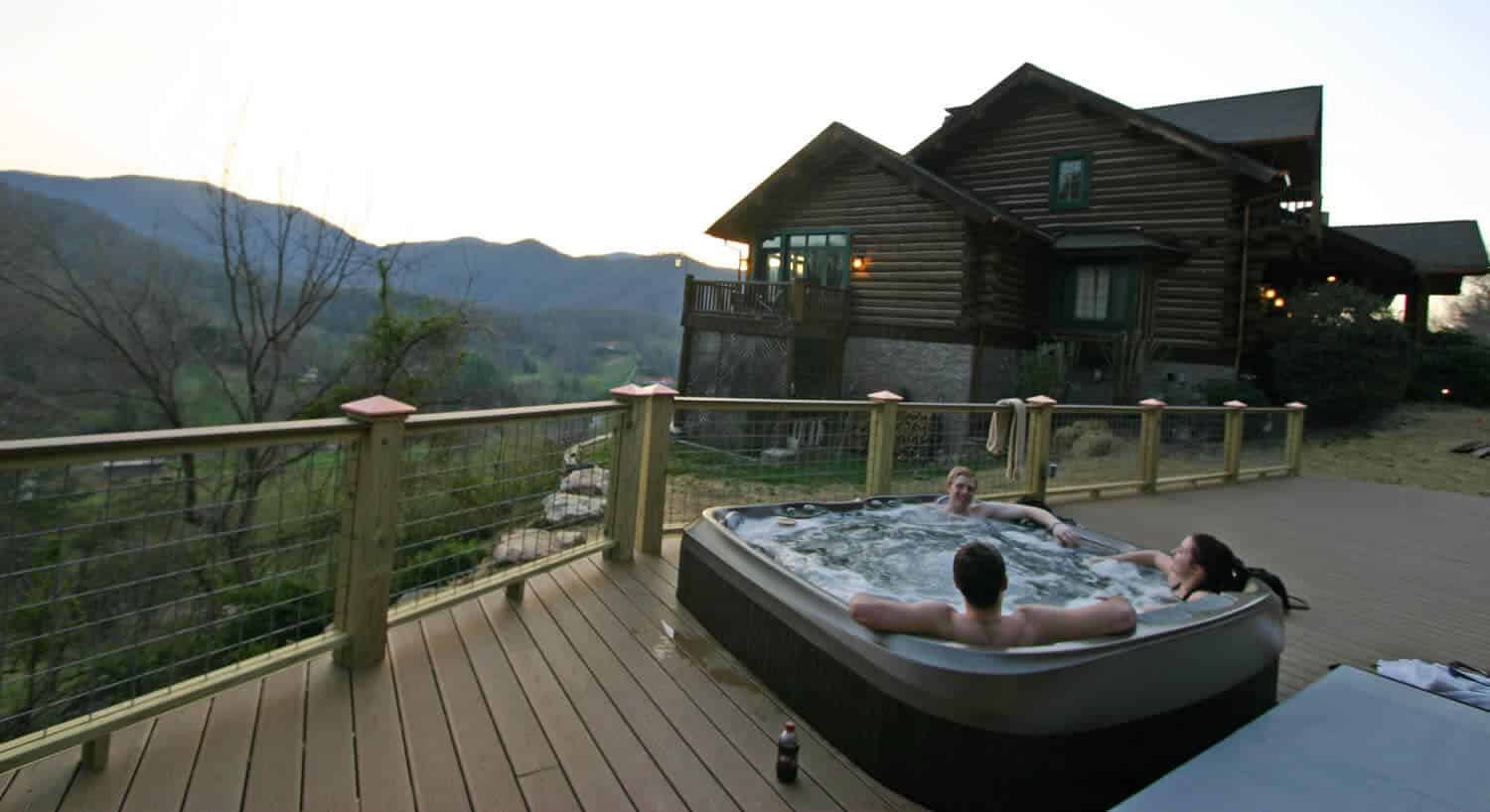 Three people enjoy a hot tub on a wooden deck next to a large log-cabin style home.