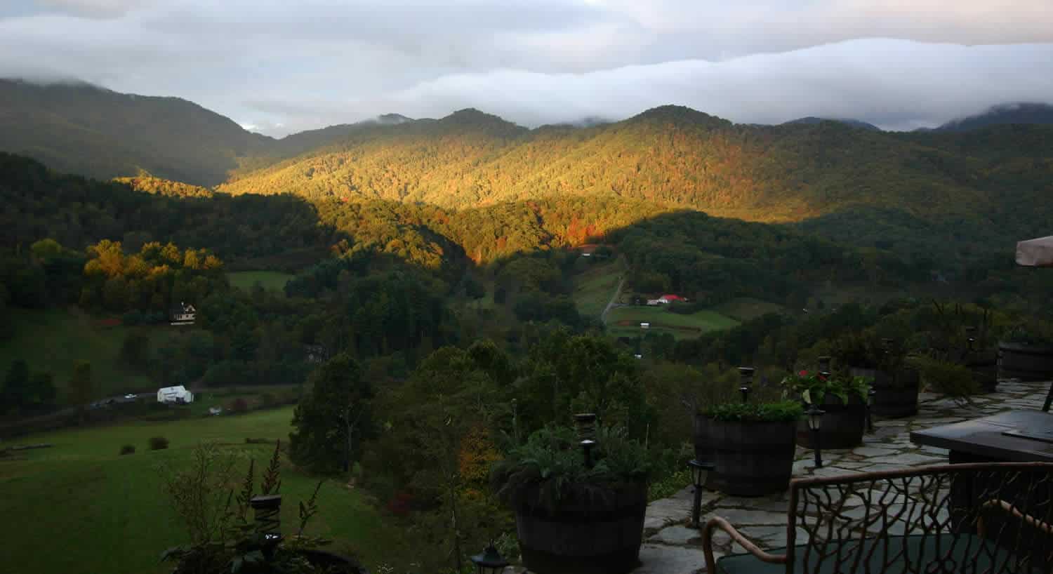 The sun rises over the mountain valley, with wooded hills surrounding a stone patio. 
