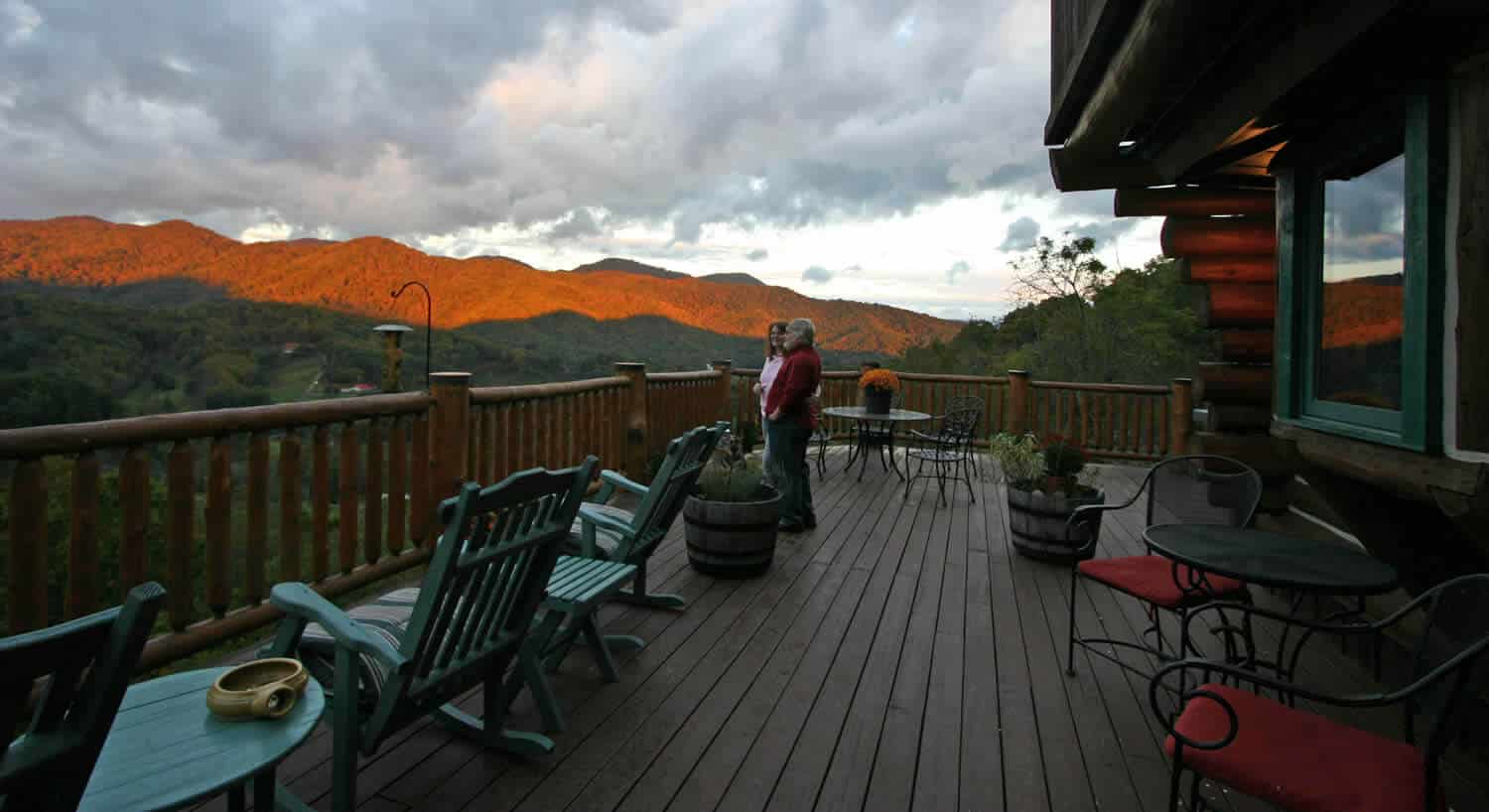Couple standing together on a wooden deck with green rocking chairs overlooking a mountain valley. 