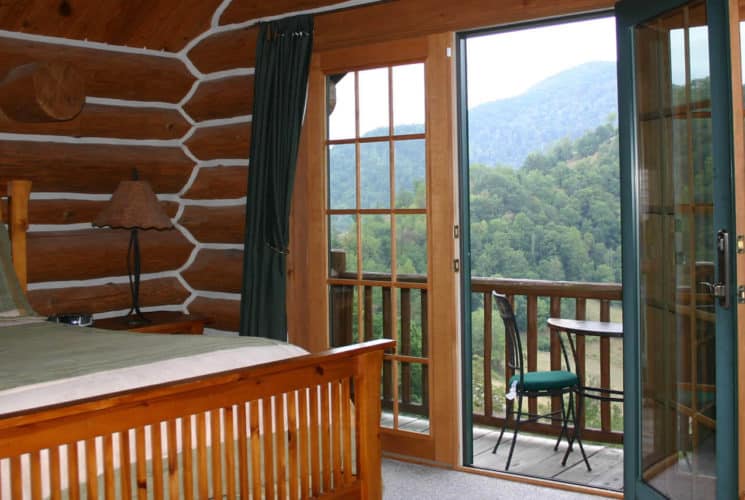 log-cabi bedroom with bed next to private balcony overlooking wooded hills.