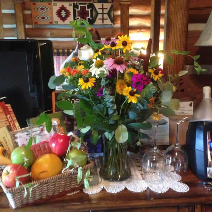 Pretty wildflowers in a glass vase next to a basket of fruit and snacks.