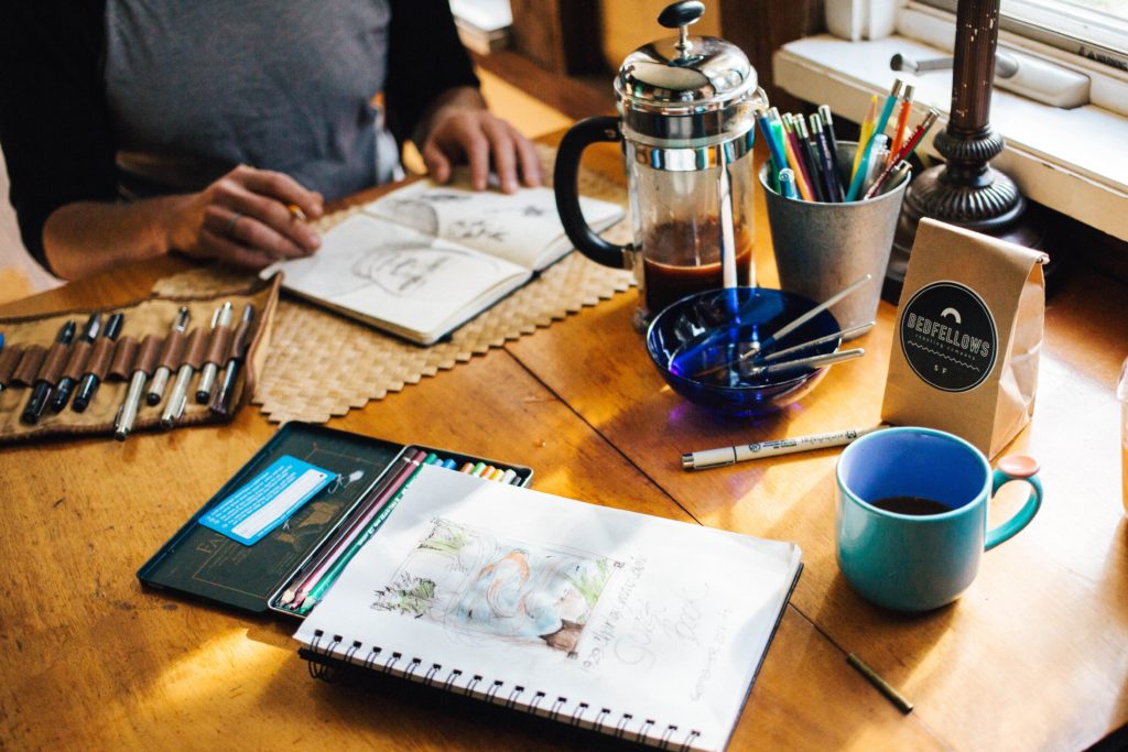 Torso of man at wood table with a sketchpad in front of him and another across from him, pencil in hand. Also on table: Pencils in roll-up holder and cup, french press coffee pot, coffee cup.