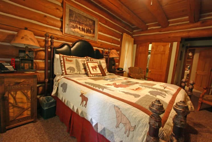 Log-cabin bedroom with large bed made up in a woods-themed quilt, two chairs and nightstands with lamps.