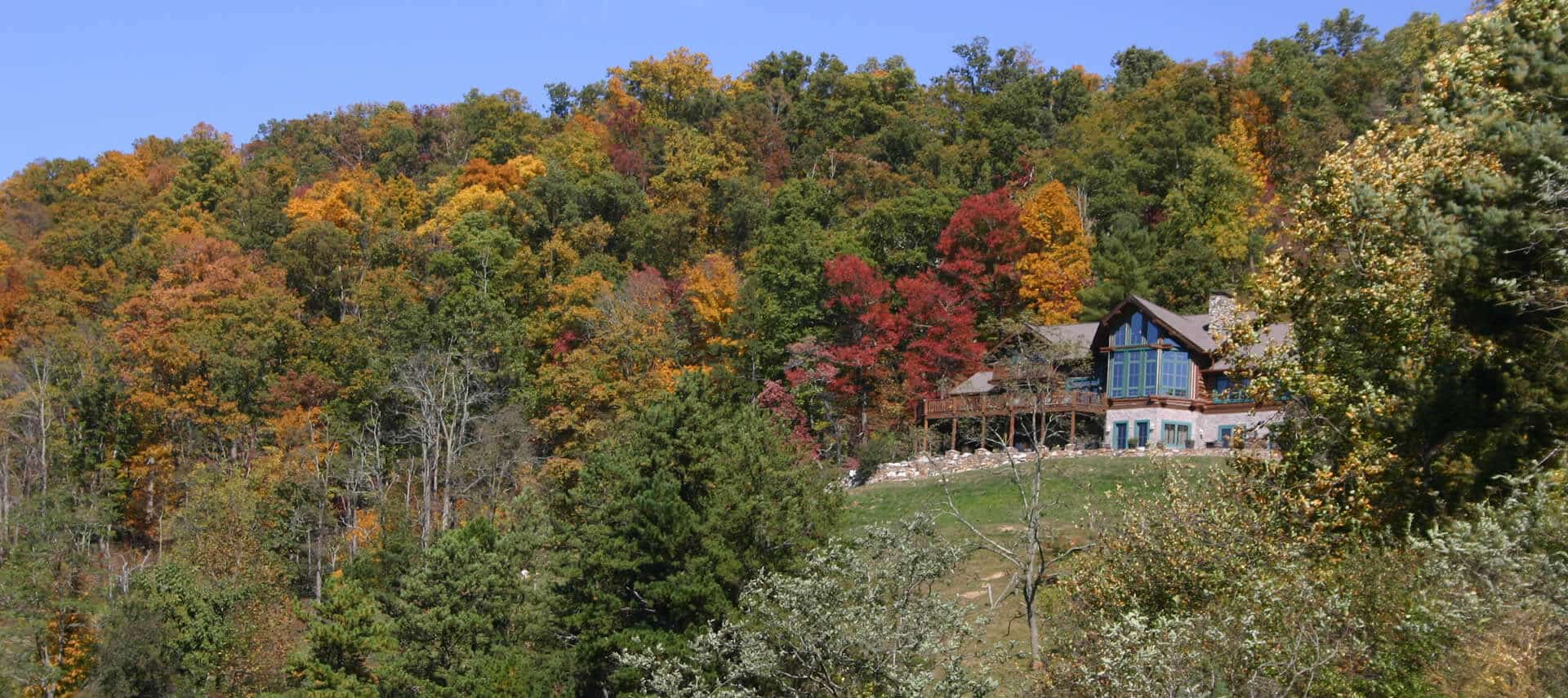 Large log-cabin style home with wooden deck nestled among hills covered in fall foliage.