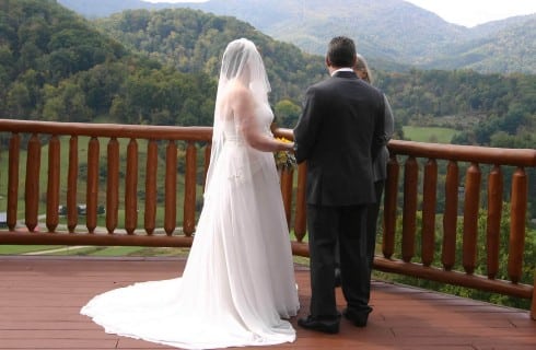 Bride in long flowing white gown with train and vail and groom in dark gray suit on wood deck facing the mountains exchanging vows