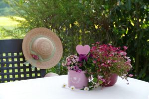 Outside table with 2 pots of pink flowers. Grid-back chair with ribboned straw hat hanging on it. Bush behind it.