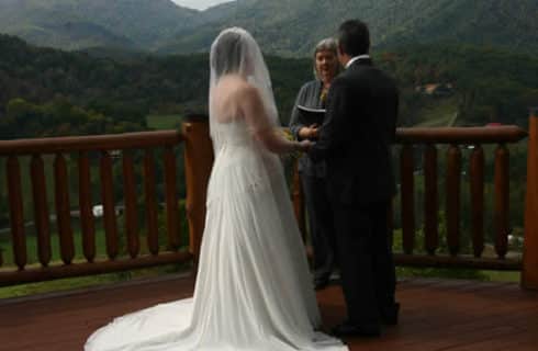 A bride in a white gown and grrom in a black tuxedo say their vows on a deck overlooking wooded hills.