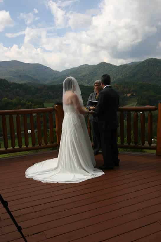 A bride in a white gown and grrom in a black tuxedo say their vows on a deck overlooking wooded hills.