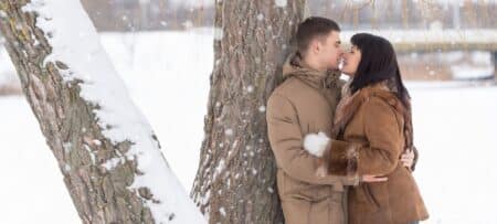 man and woman face to face in each other’s arms leaning against a snow-cover tree trunk