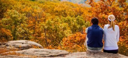Couple sitting side side on a rocky ledge surrounded by colorful trees during the fall