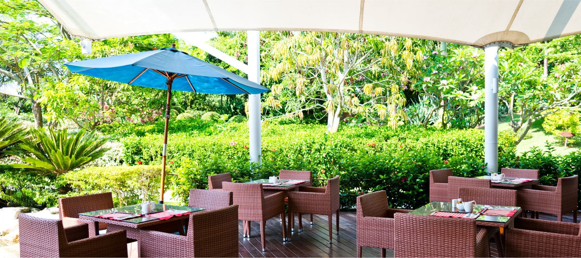 A group of tables on a green outdoor patio, one with a blue umbrella and a cream sail cover over all the tables.